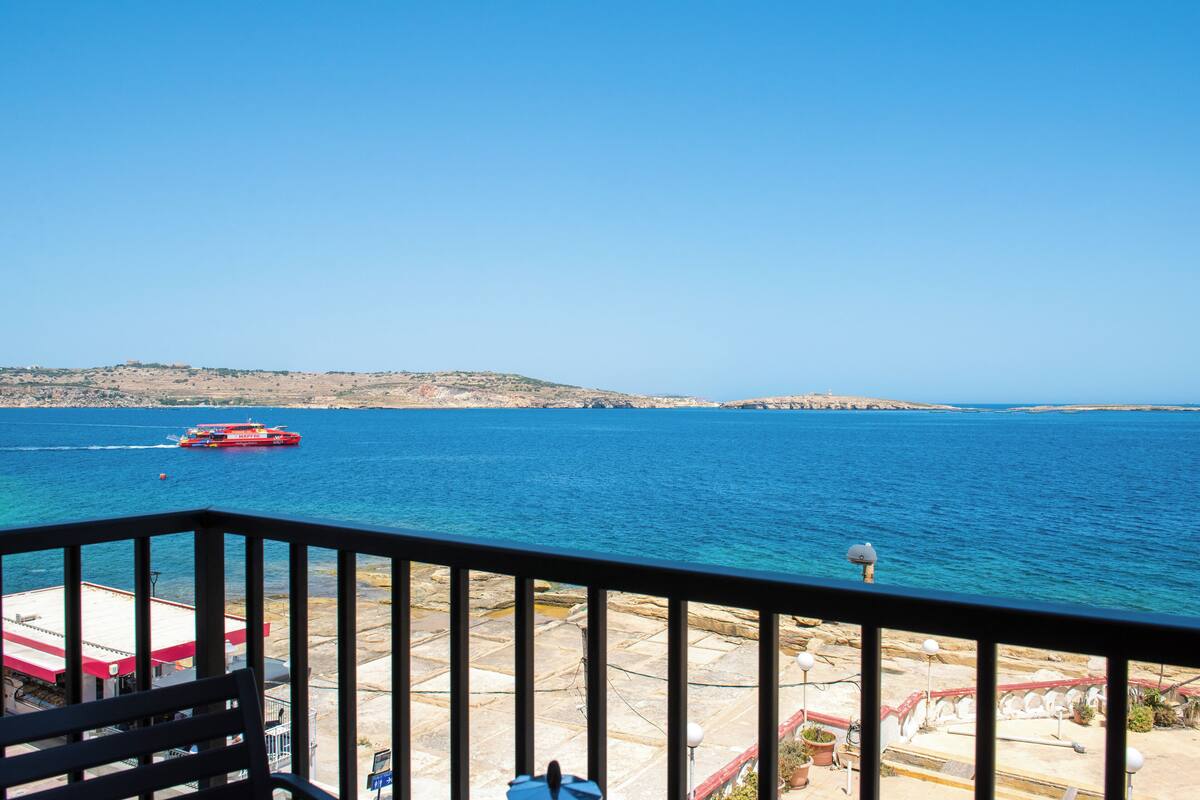 A balcony with a view of the sea, a red boat in the distance, and rocky terrain across the water.