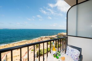 A balcony with a view of the sea and beach, a bottle of champagne, and a table with a glass and a plate. - Islet Seafront Family Apartment  (St. Paul's Bay)