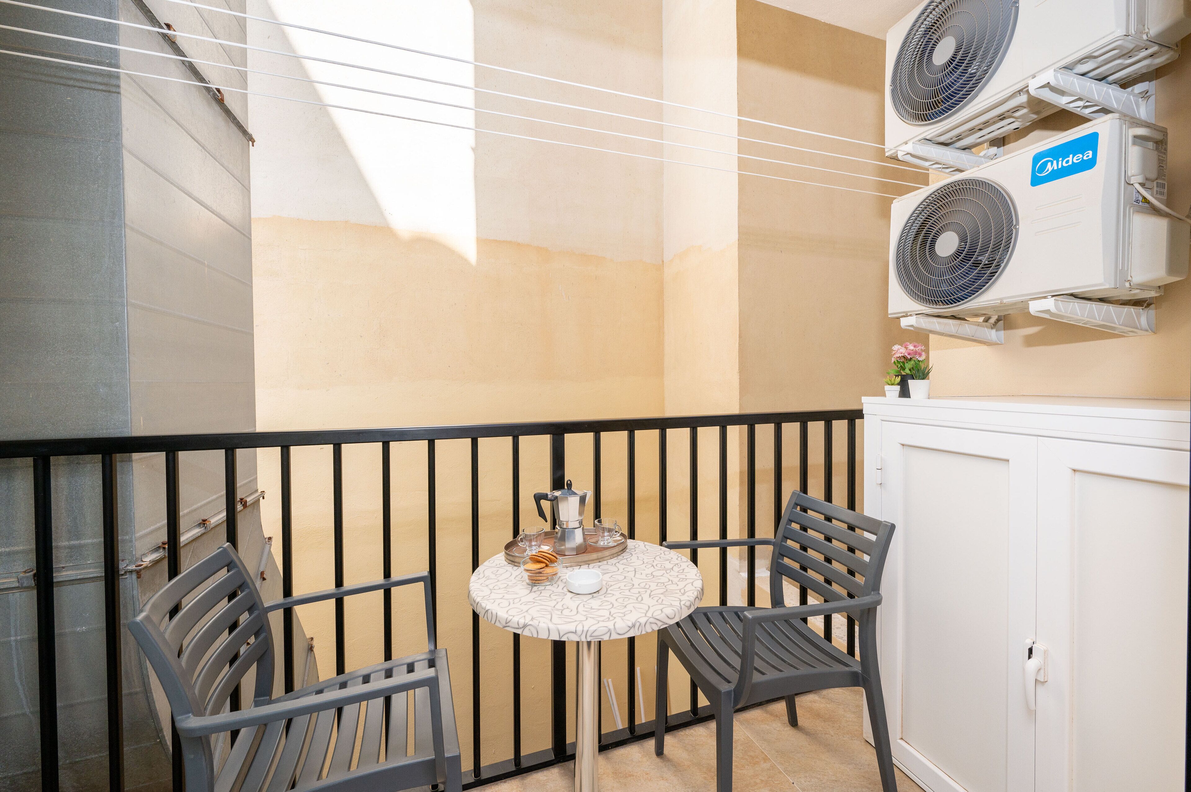 A balcony with a table, two chairs, and a cabinet. There are two air conditioning units mounted on the wall.