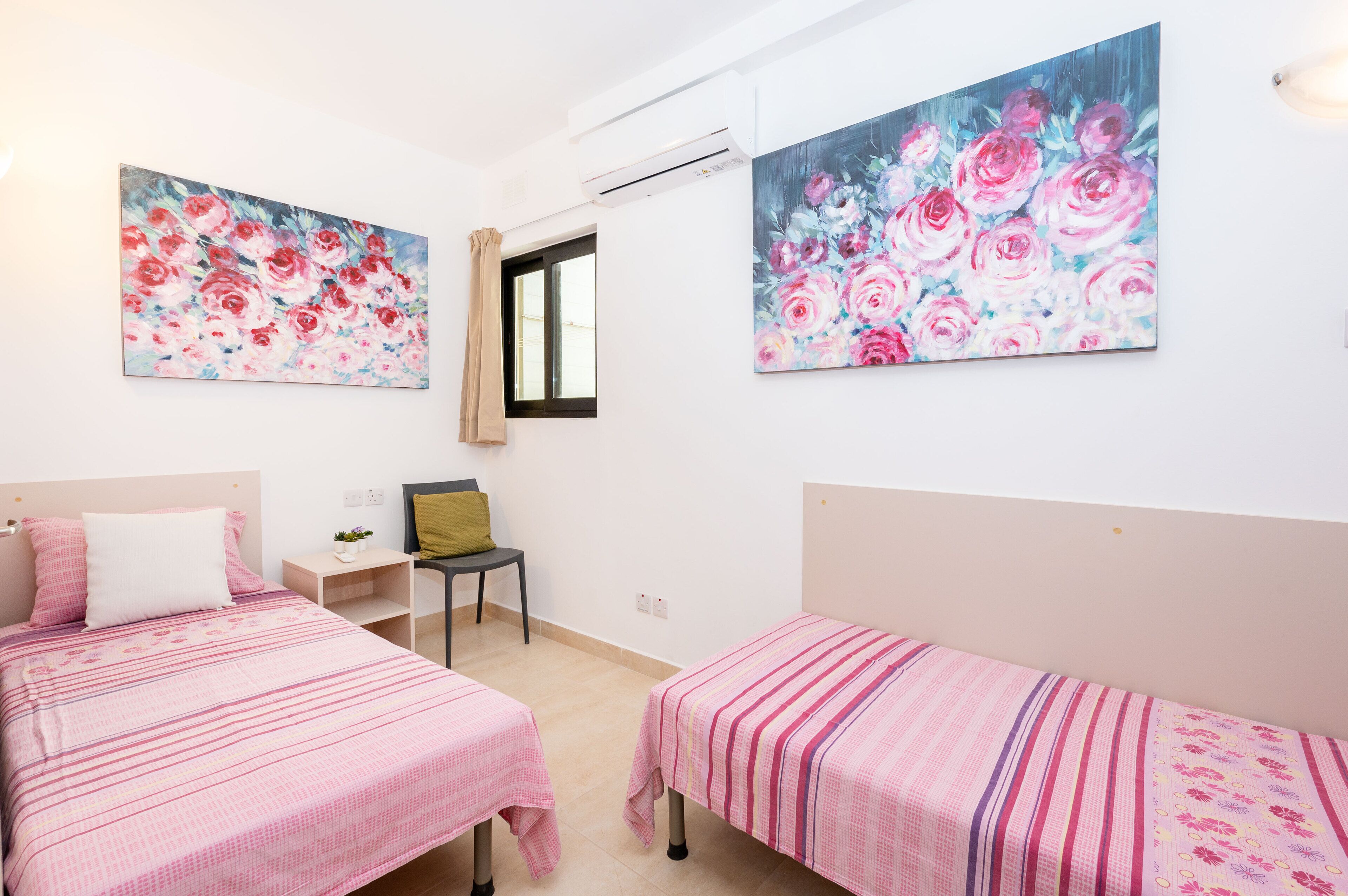 A bedroom with two beds, a desk, and a chair. There are two floral paintings on the walls.