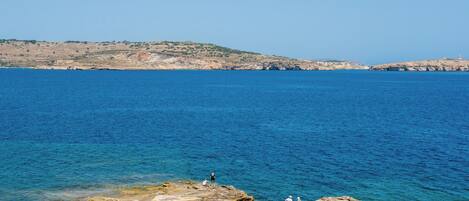 Vista costera con mar azul cristalino, costa rocosa y colinas al fondo.