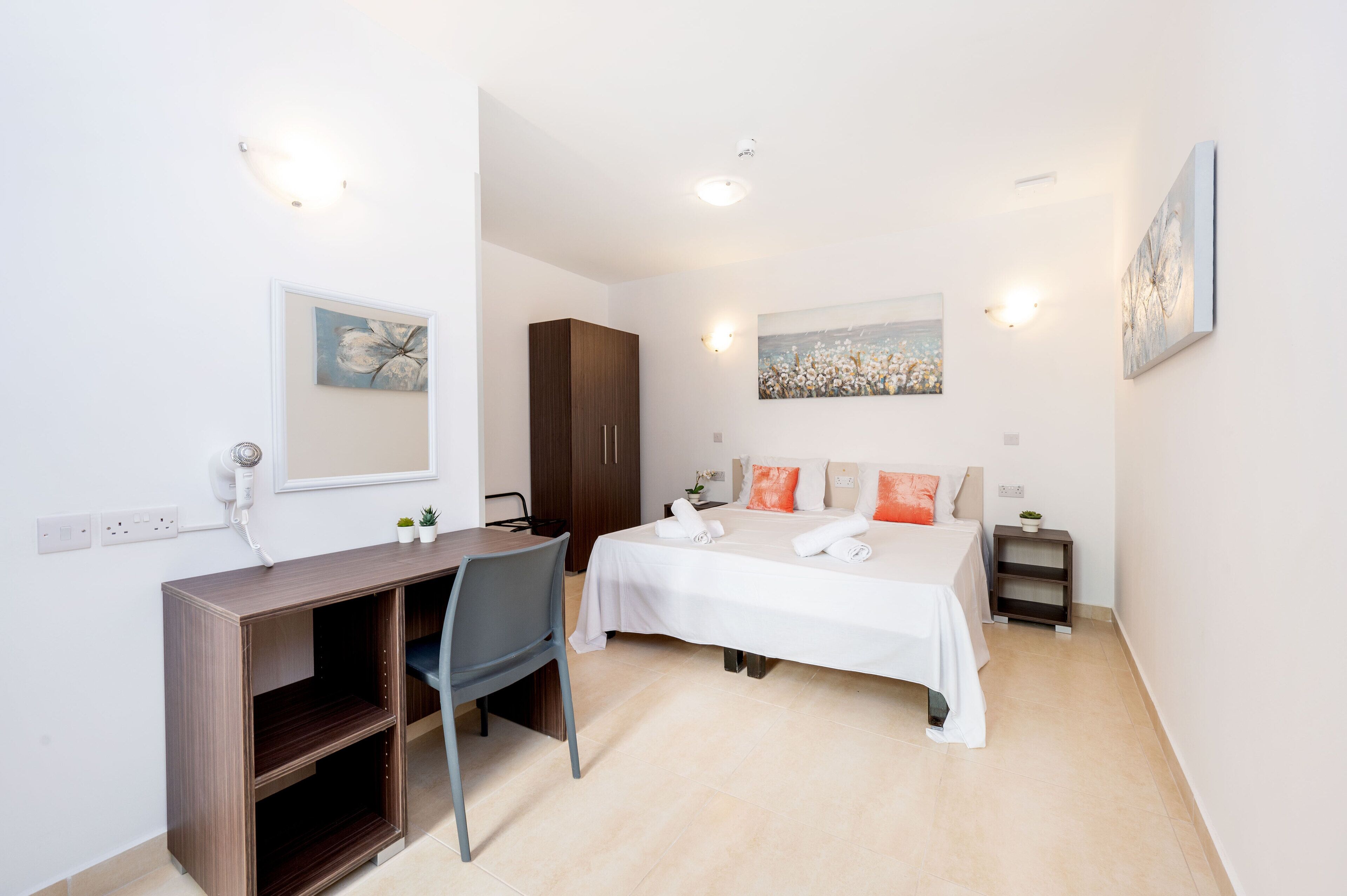 A hotel room with a bed, a desk, a chair, a wardrobe, and a small bedside table with a lamp.