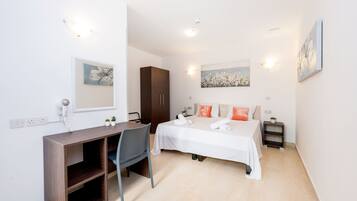 A hotel room with a bed, a desk, a chair, a wardrobe, and a small bedside table with a lamp.