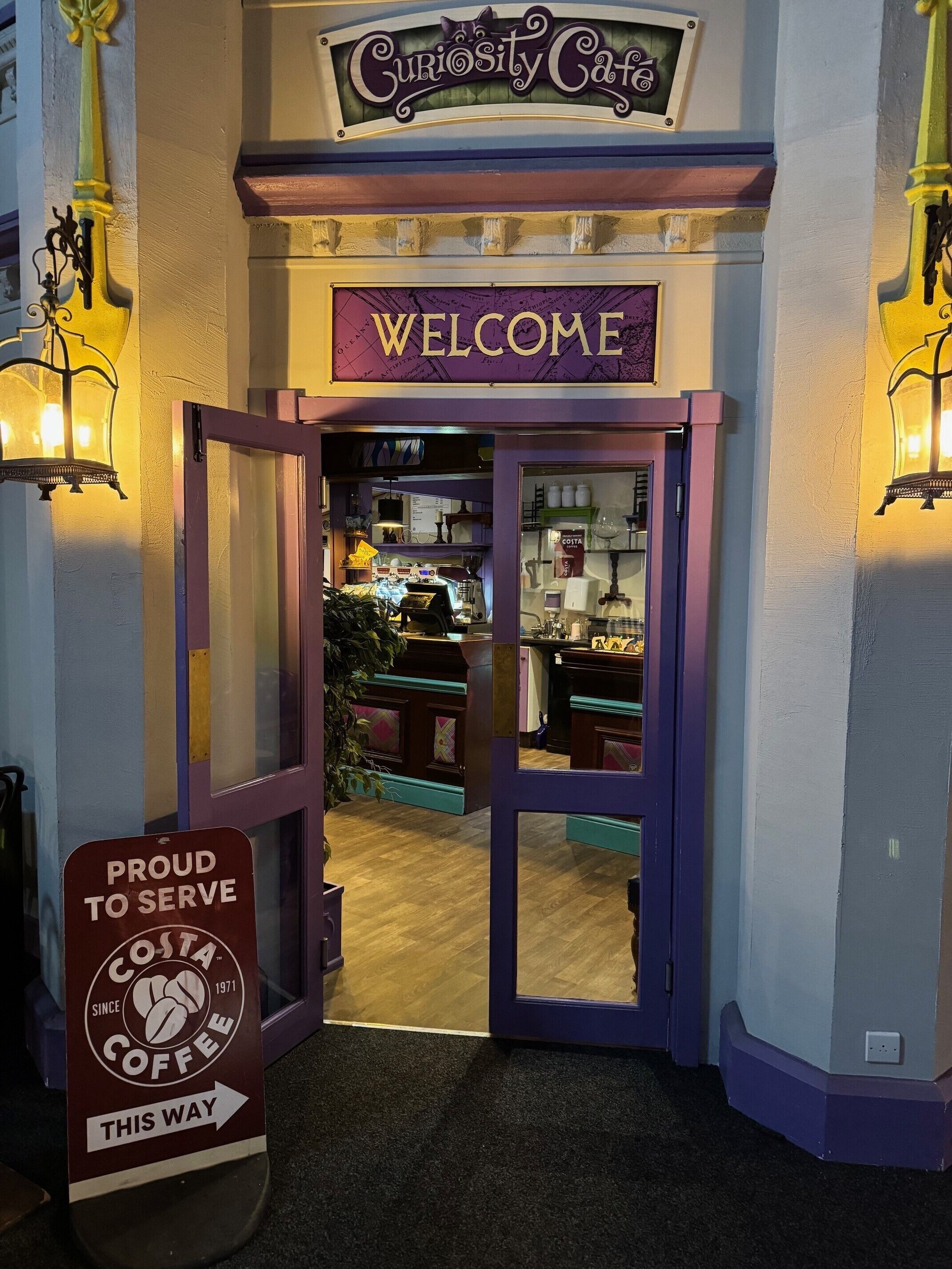 A coffee shop entrance with a 'Welcome' sign, a 'Proud to Serve Costa Coffee' sign, and a 'Curiosity Cafe' sign above.