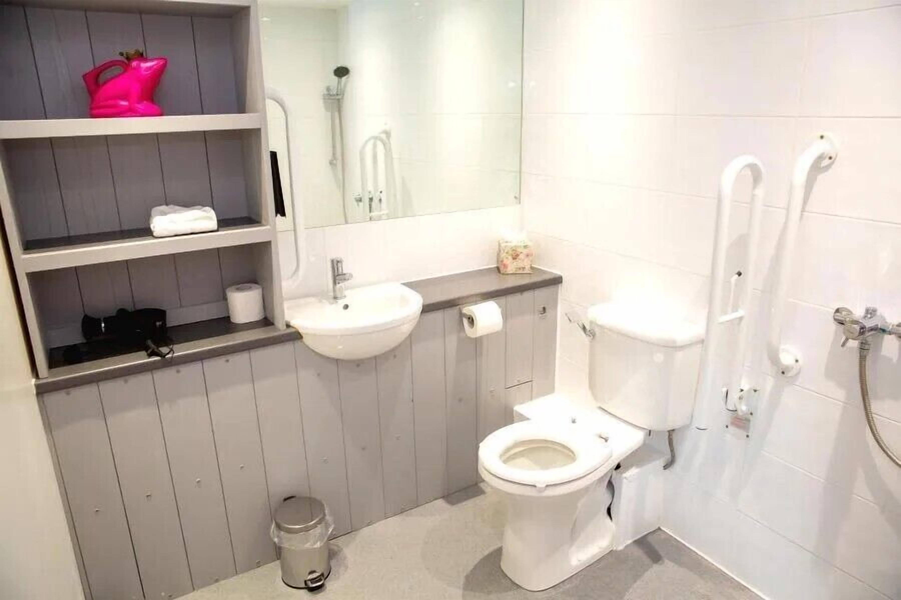 A bathroom with a toilet, sink, and shower. There are shelves with toiletries and a mirror above the sink.