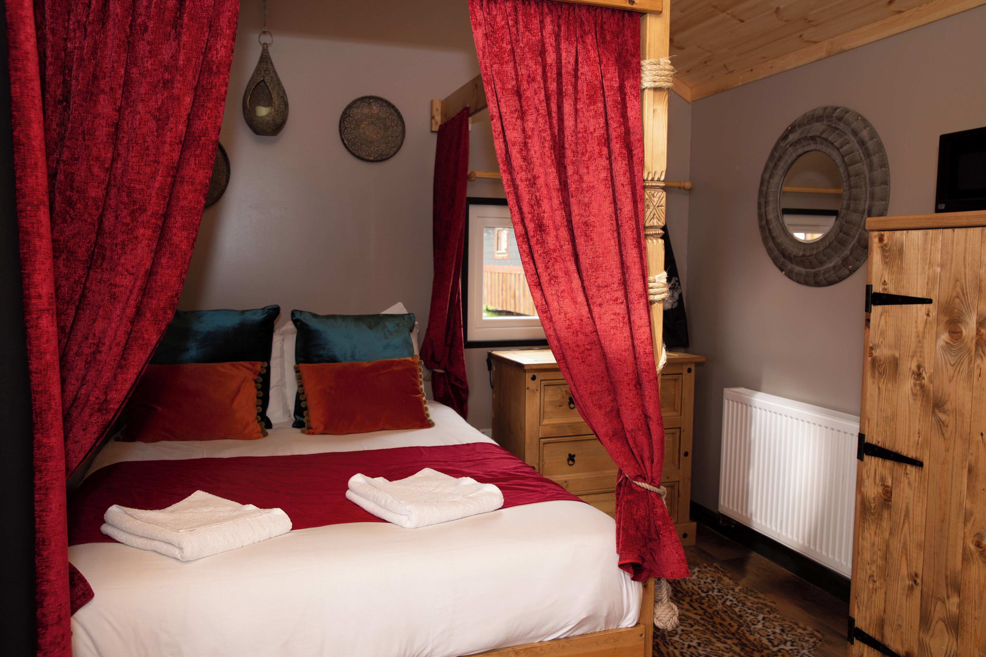 A bedroom with a bed, red curtains, a wooden wardrobe, a mirror, and a radiator.