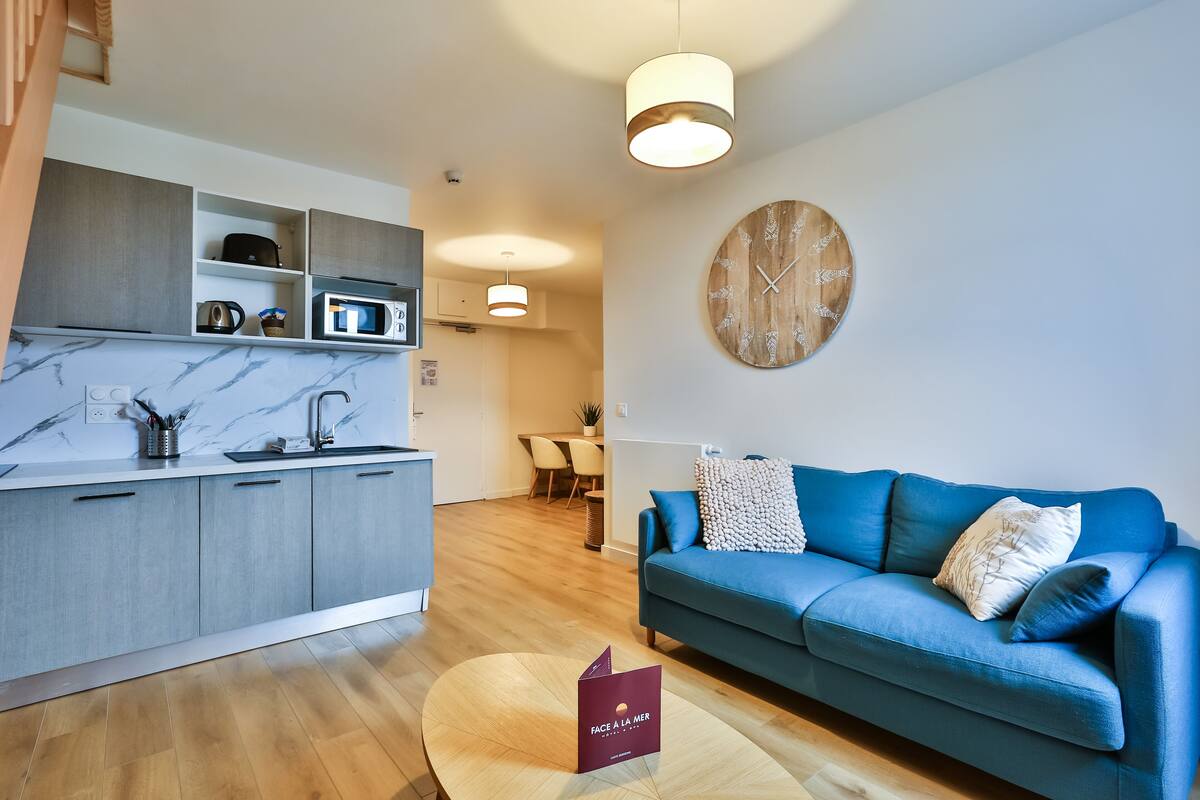 A modern living room with a blue sofa, a round wooden clock, and a kitchen area with grey cabinets and a marble countertop.