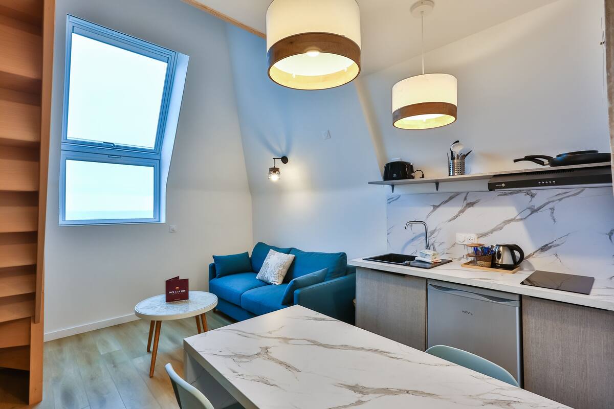 A compact kitchen with a built-in oven, a coffee machine, and a microwave. There is a blue sofa and a small round table with a red folder.
