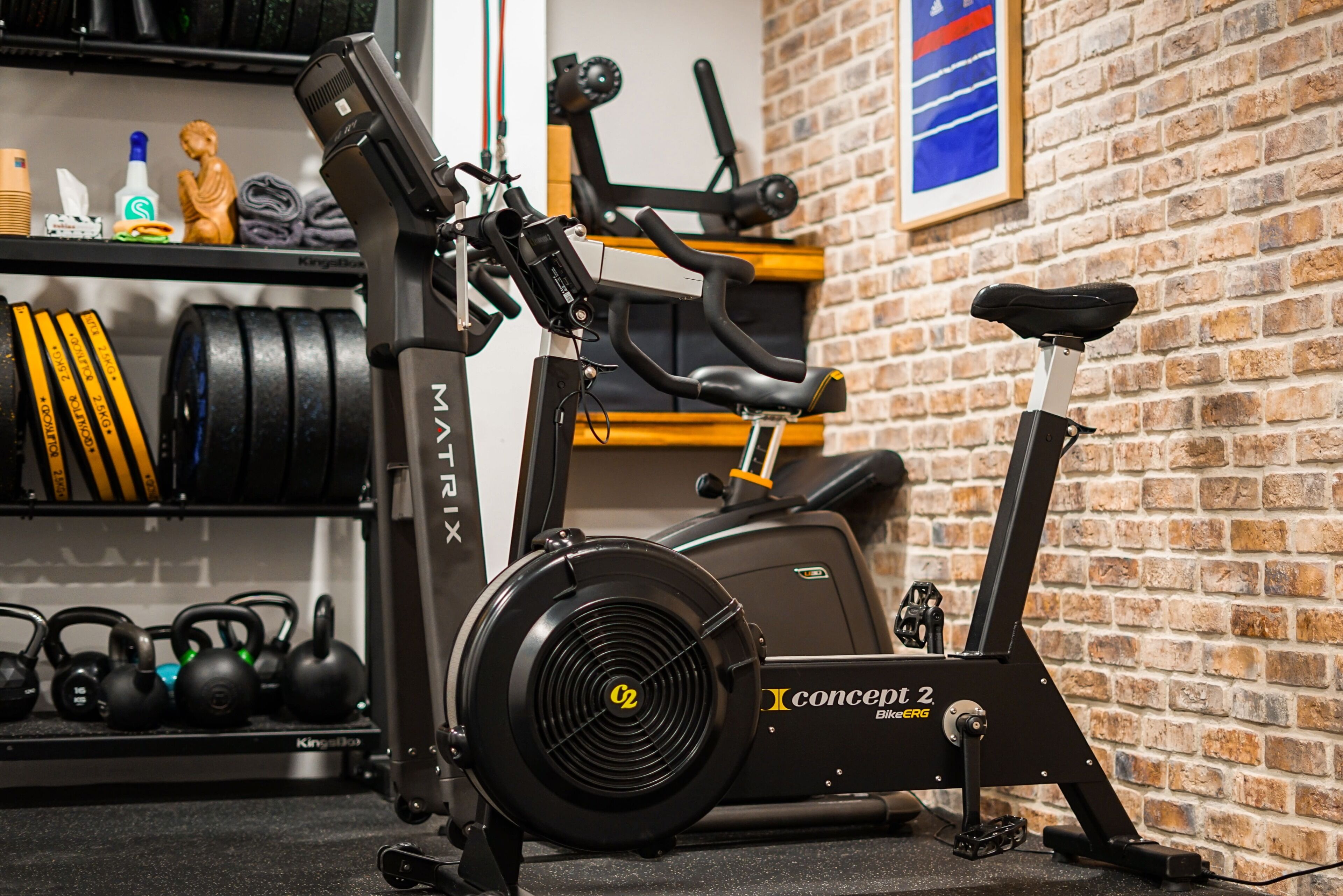A stationary bike in a room with a brick wall and a shelf with weights and kettlebells.