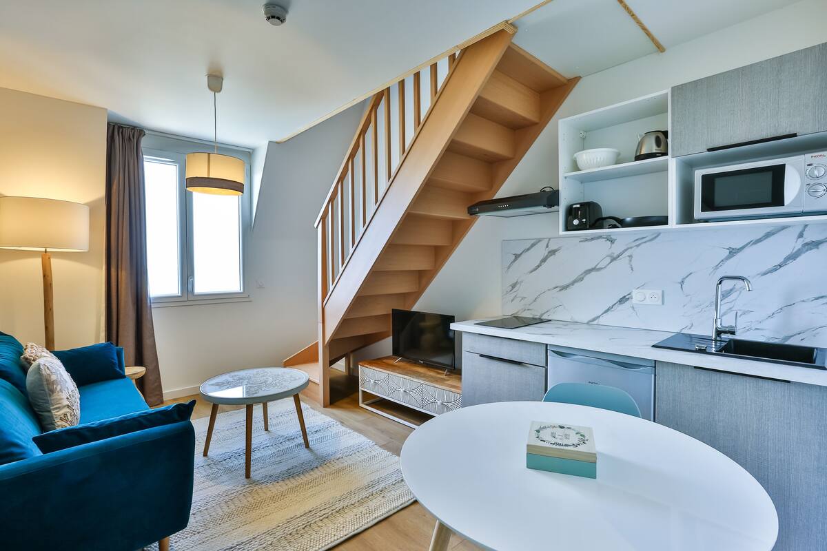 A modern living room with a blue sofa, a small round table, and a staircase leading to a kitchen area with a microwave and cabinets.