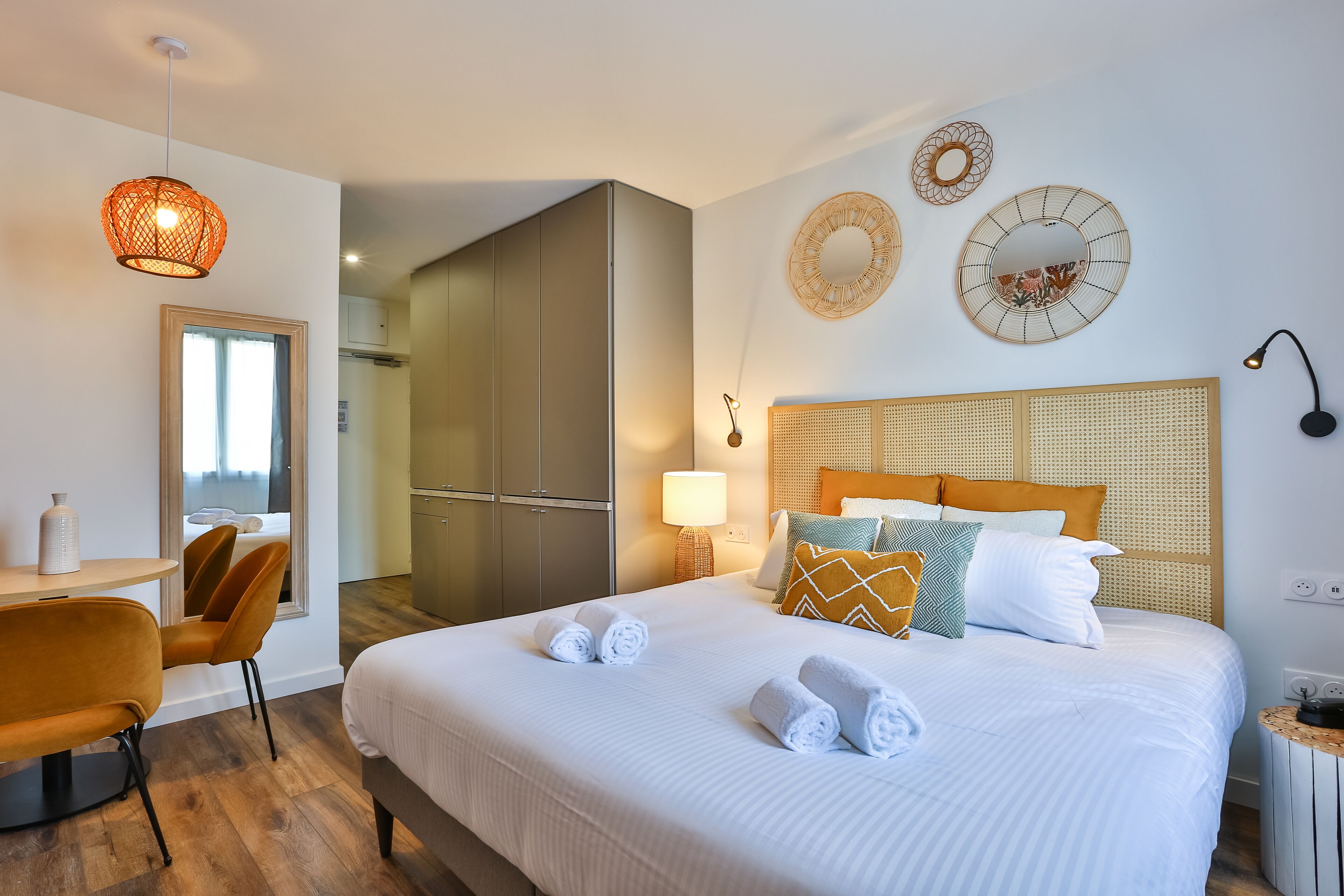 A modern hotel room with a large bed, a dining area with a table and chairs, and a view of the city through a window.