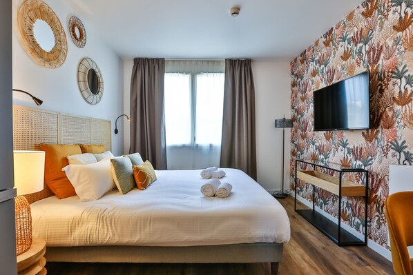 A hotel room with a large bed, a flat-screen TV, a wooden side table, and a wall with a floral wallpaper.