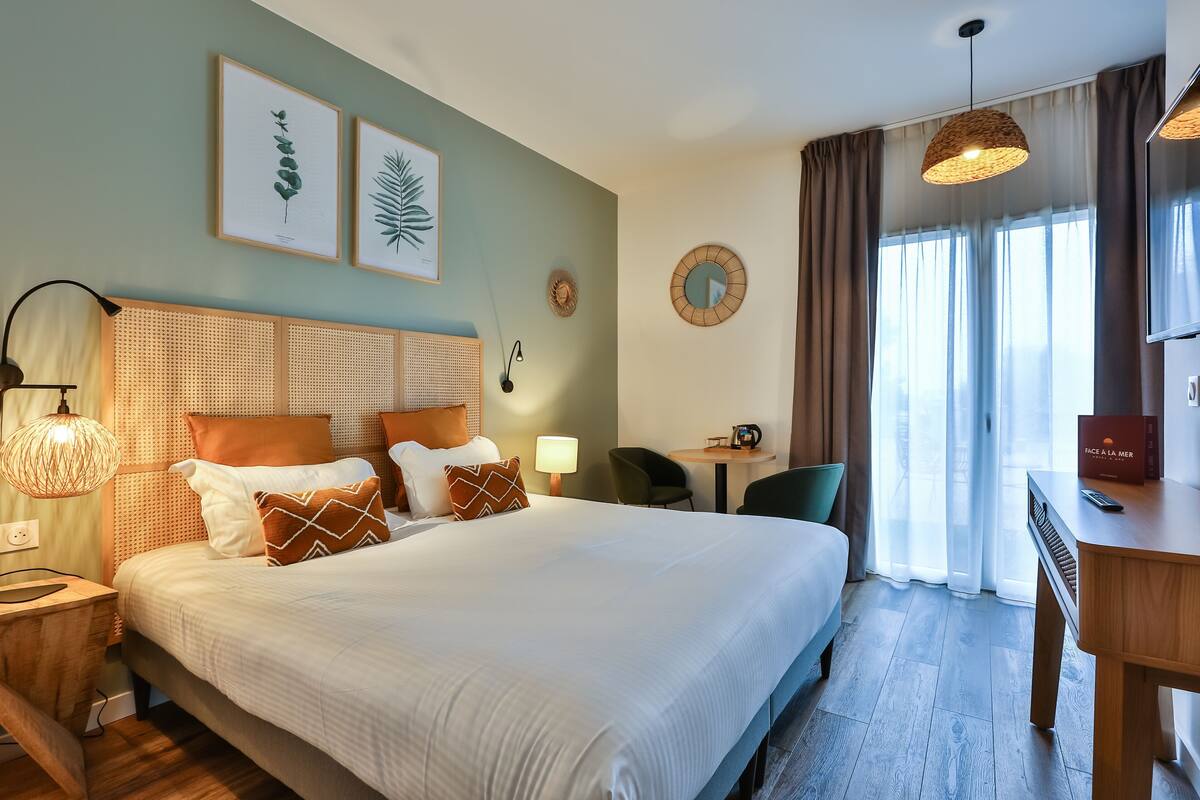 A hotel room with a bed, bedside table, lamp, framed artwork, and a view of the outdoors.
