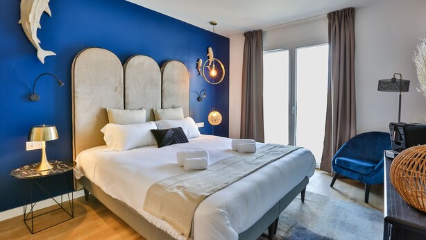 A modern bedroom with a large bed, a blue accent wall, and a large window. - Hôtel Spa Face à la Mer (Merville-Franceville-Plage)