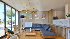 A modern interior with a large sectional sofa, wooden chairs, a coffee table, and a reception desk with a marble countertop. - Hôtel Spa Face à la Mer (Merville-Franceville-Plage)