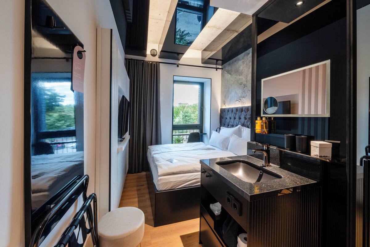 A modern hotel room with a bed, a large window, a bathroom with a sink and mirror, and a TV mounted on the wall.