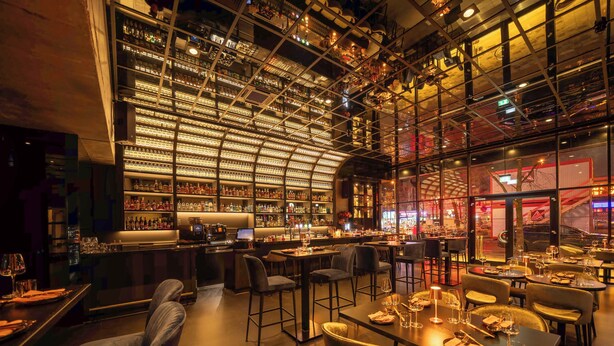 A bar area with a high ceiling and shelves stocked with bottles. There are tables and chairs for dining.