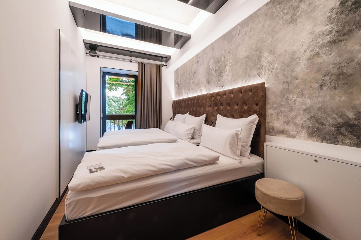 A modern bedroom with a large bed, a small stool, and a view of greenery outside.