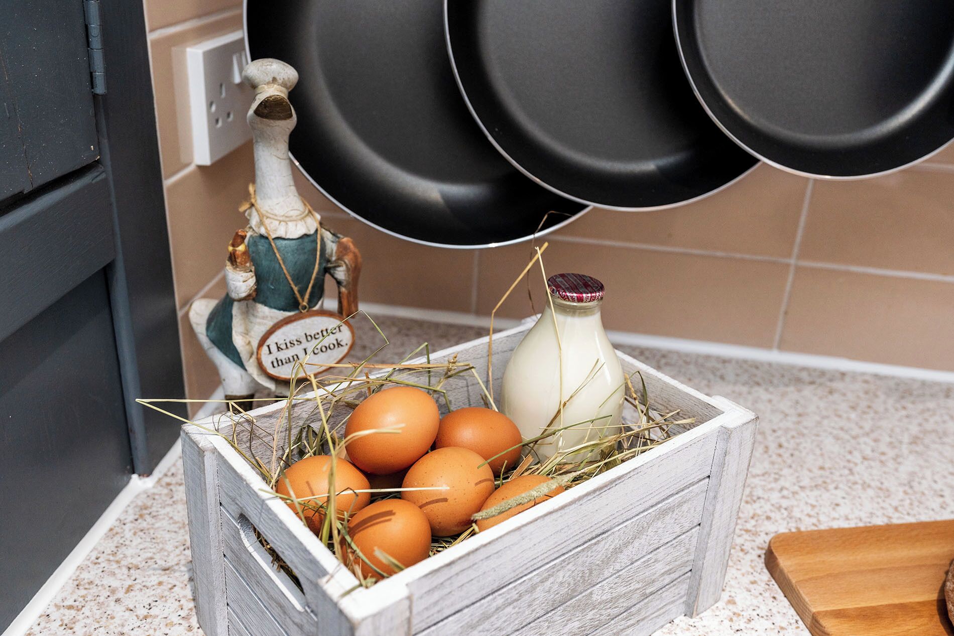A kitchen counter with a wooden crate containing eggs and a bottle of milk, a decorative figure, and hanging black pans.
