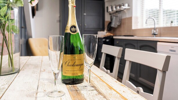 En flaska champagne och två glas på ett träbord i ett kök med en vit stol och en vas med blommor.