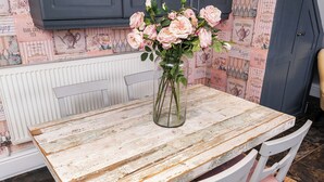 Ett rustikt träbord med en vas med rosa rosor, vita stolar och tapet med ett motiv som för tankarna till te och en svunnen tid.