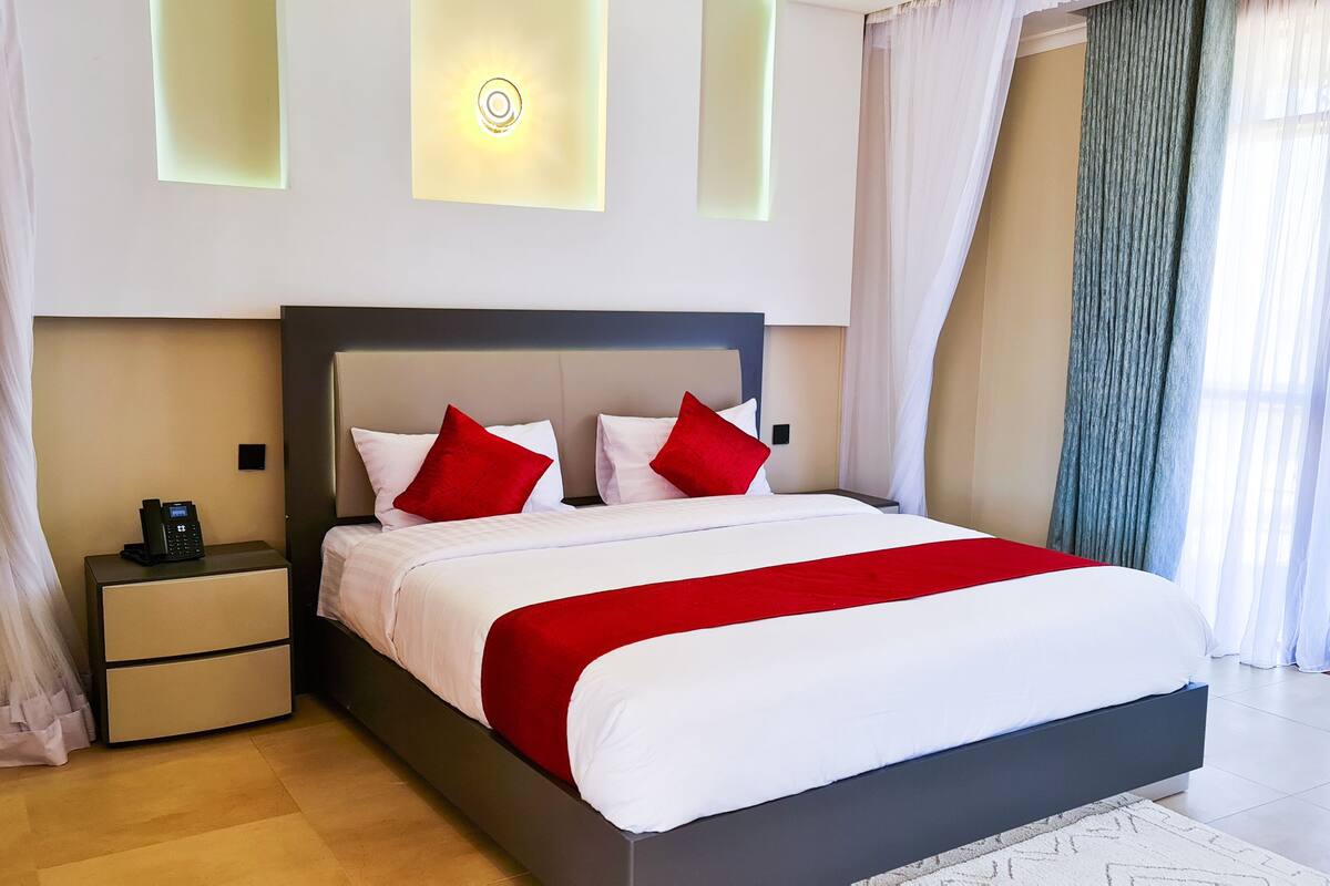 Una habitación de hotel moderna con una cama grande, ropa de cama blanca, almohadas rojas, un teléfono en la mesita de noche y una mesita de noche.