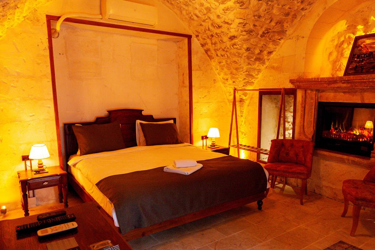 A bedroom with a four-poster bed, a fireplace, a red armchair, and a nightstand with a lamp.