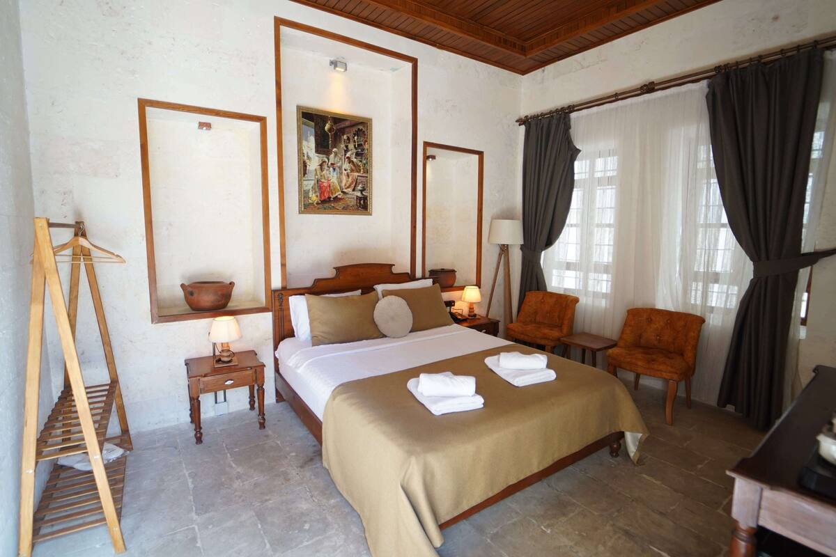 A hotel room with a bed, two chairs, a desk, a lamp, a painting, and a wooden ladder.