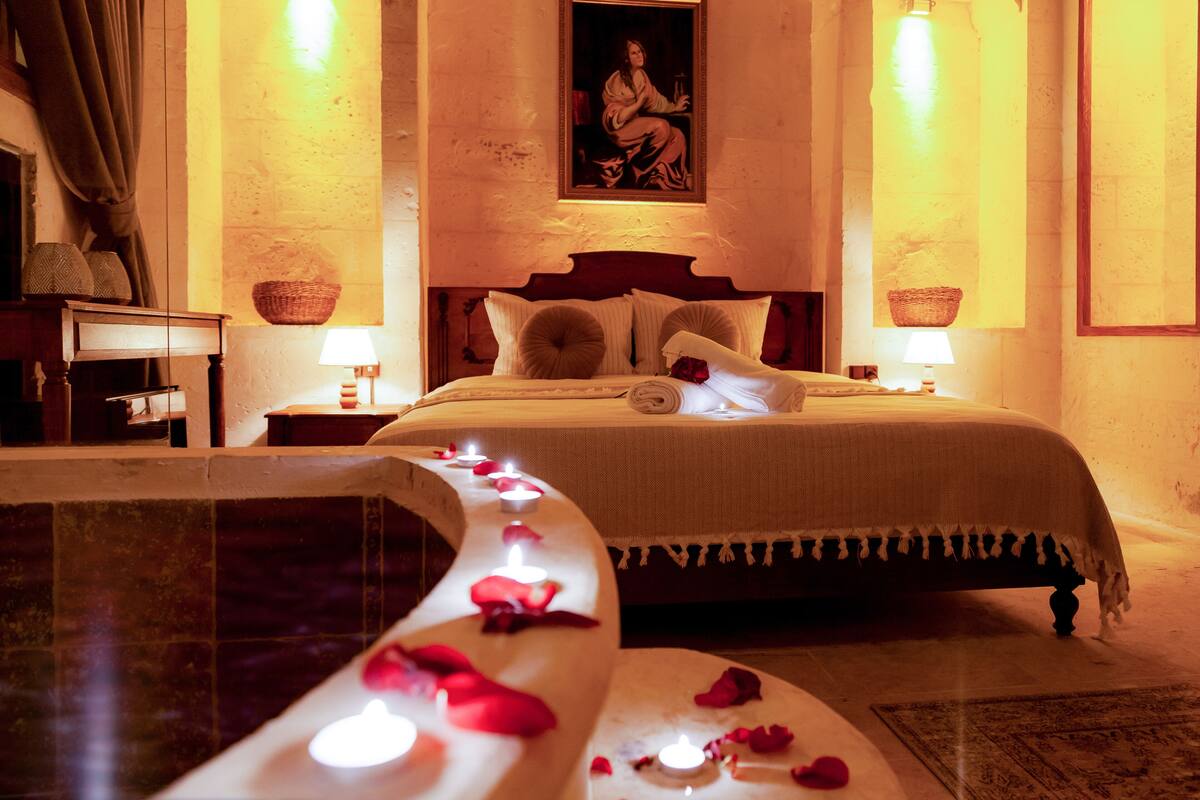 A cozy bedroom with a bed, a nightstand, a painting, and a bathtub with rose petals and candles.