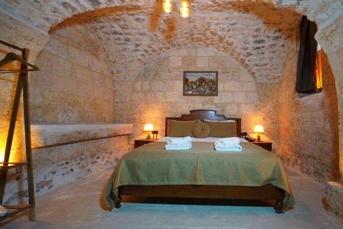 A cozy room with a stone wall, a bed with a green bedspread, two bedside tables with lamps, and a painting on the wall.