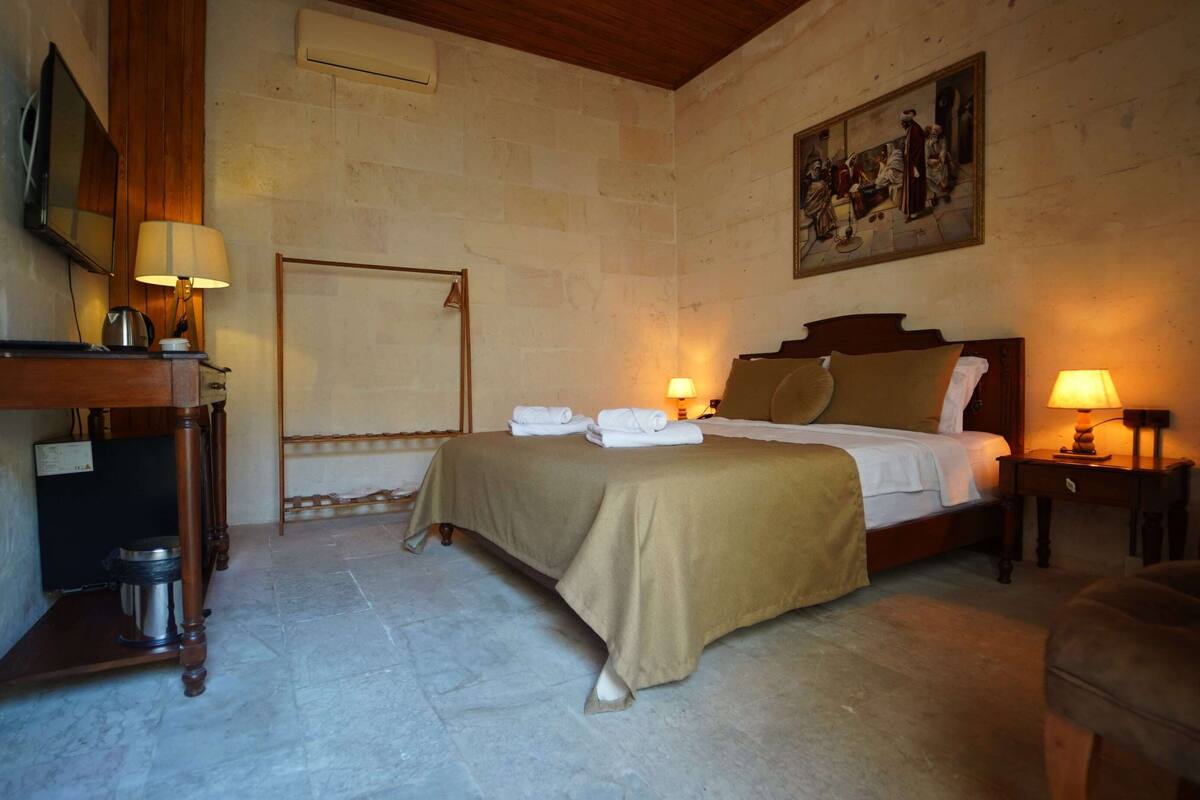 A hotel room with a bed, bedside tables, a desk, a chair, and a painting on the wall.