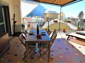 A covered outdoor dining area with a table and chairs, potted plants, and a view of buildings and mountains in the background. - Casa Alma (Positano)
