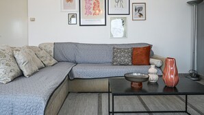 Una sala moderna con un sofá gris, cojines estampados, una mesa de centro con objetos decorativos y arte mural que incluye una hoja de palma y diseños abstractos.