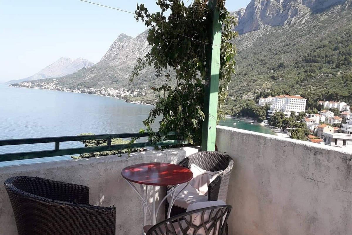 A balcony with wicker furniture and a table, overlooking a coastal town and mountains.