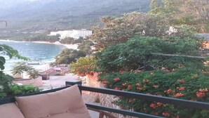 Un balcón con zona de estar acolchada que da a un paisaje costero con playa y un edificio en la distancia.