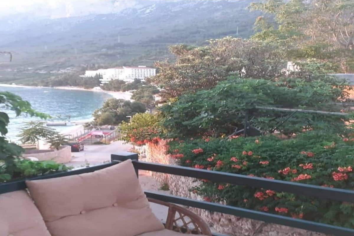 A balcony with a cushioned seating area overlooking a coastal landscape with a beach and a building in the distance.