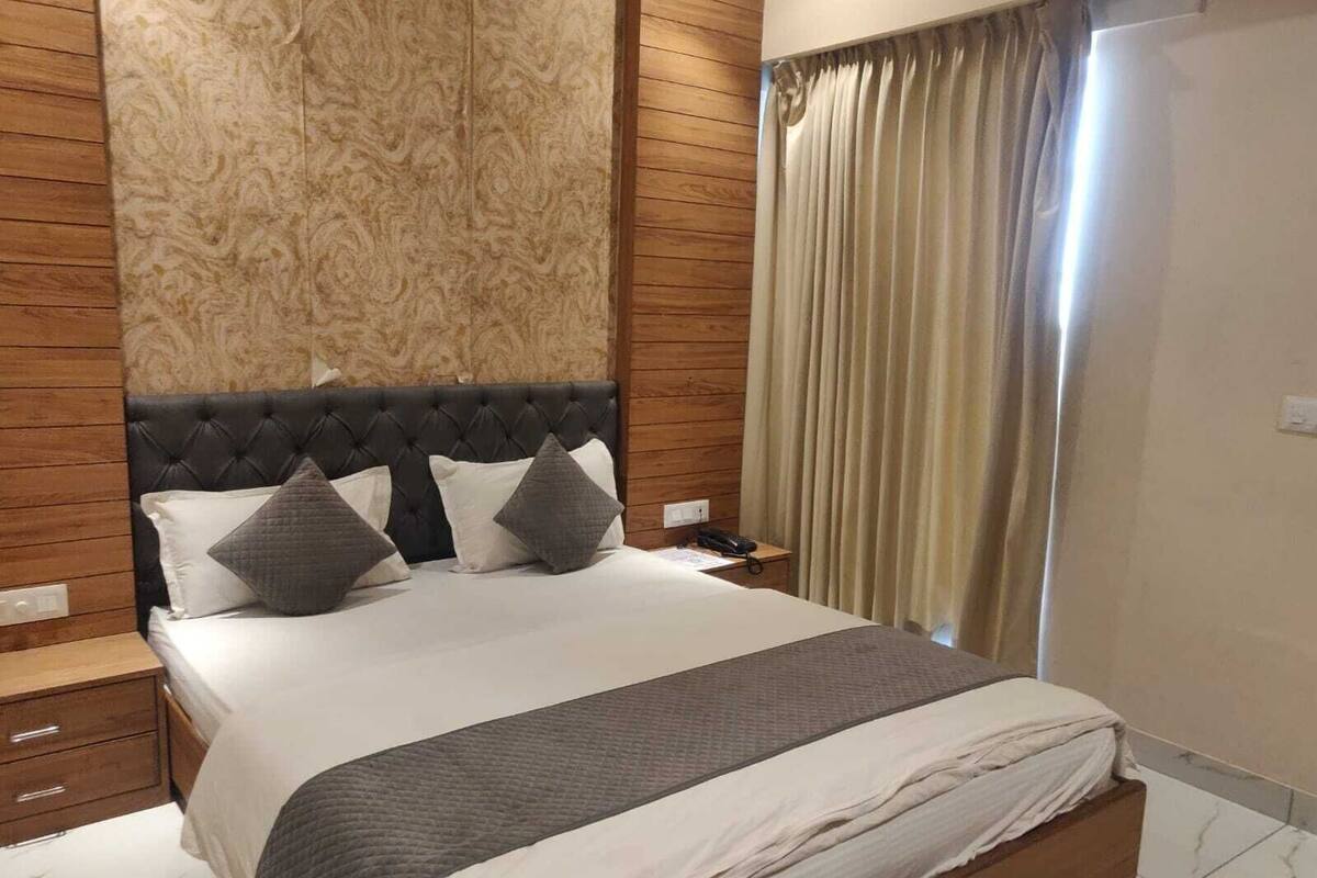 Un dormitorio con cabecera capitonada, cama con ropa de cama blanca y gris, mesita de noche de madera y una cortina.