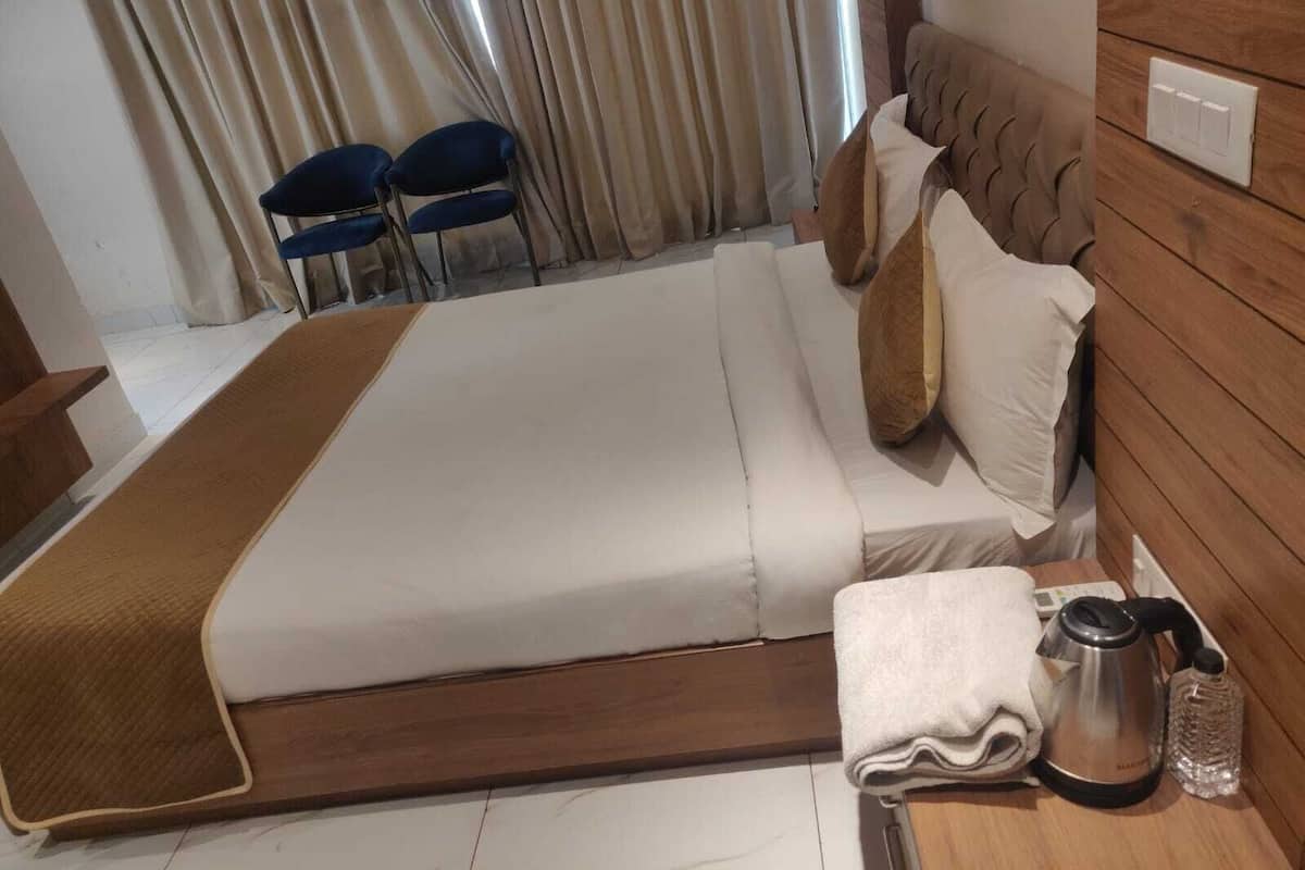 Una habitación de hotel con una cama grande, cabecera de madera, dos sillas azules, una mesita de noche de madera con una tetera, una botella y una toalla doblada.