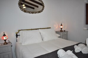 Una cama bien hecha con sábanas blancas, dos mesitas de noche con lámparas, un espejo en la pared y toallas enrolladas en el suelo.