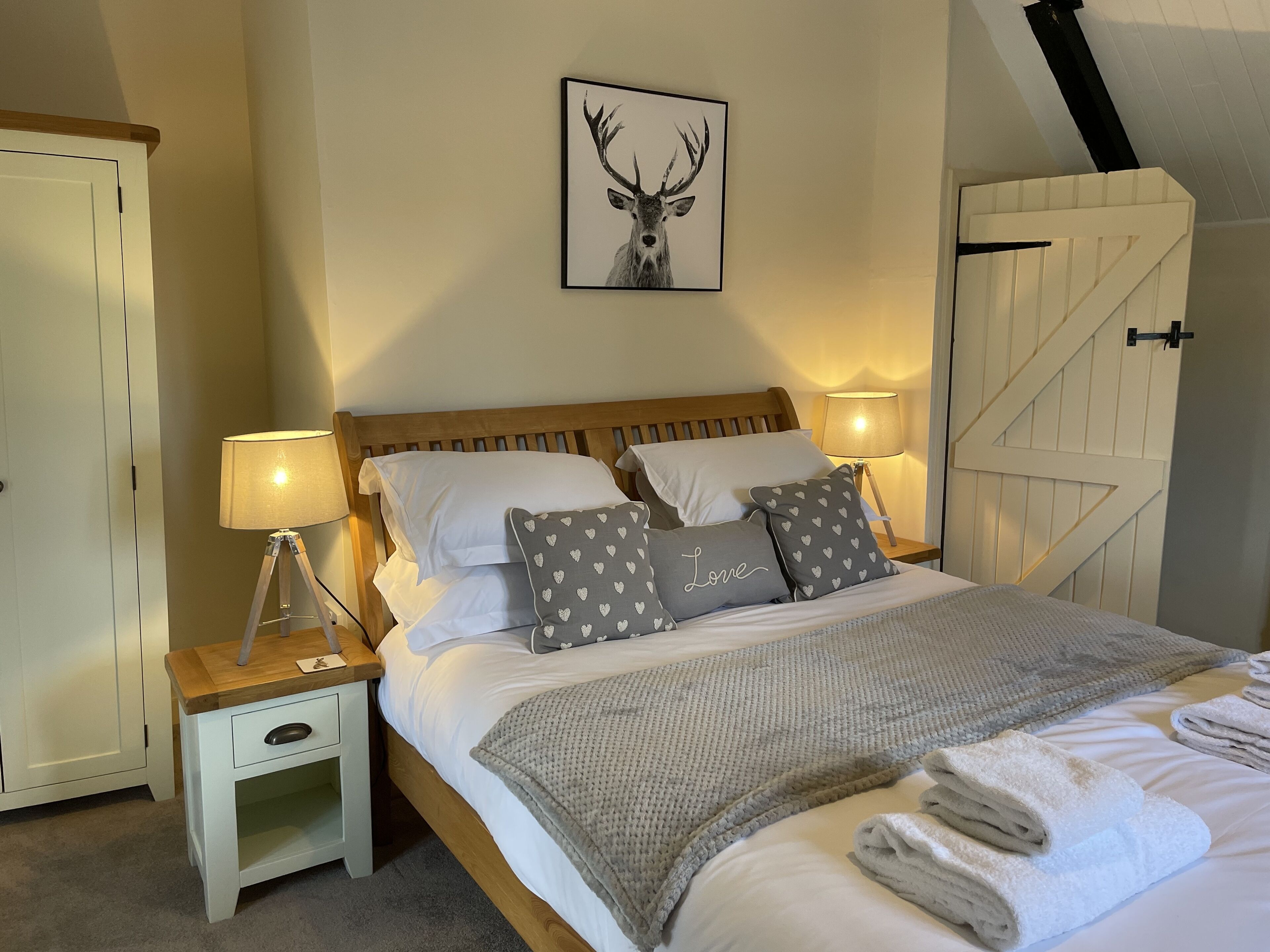 Een slaapkamer met een houten bed, witte beddengoed, kussens met een hartjesprint, een foto van een hert in een lijst, en een witte deur.