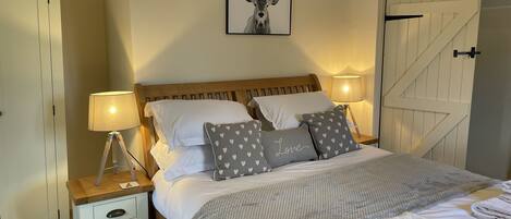Een slaapkamer met een houten bed, witte beddengoed, kussens met een hartjesprint, een foto van een hert in een lijst, en een witte deur.