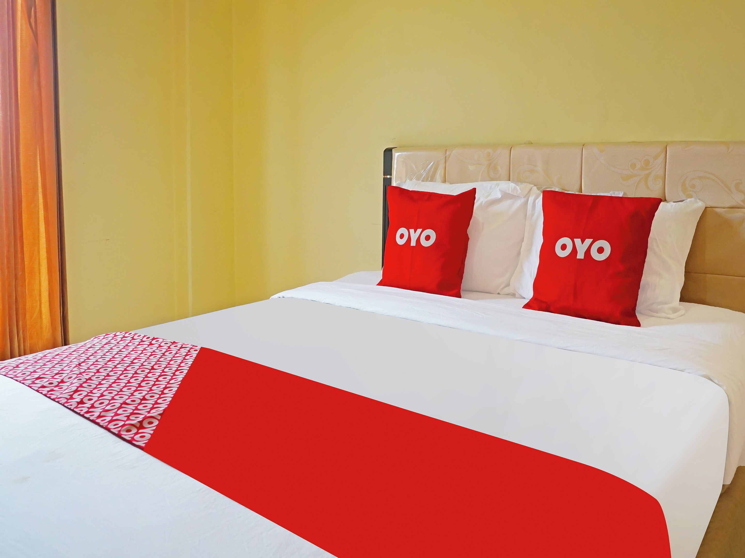 A bed with a white and red bedspread, two red pillows with the word 'OYO' on them, and a yellow wall.