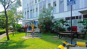A playground with swings and a slide, surrounded by greenery and a building with a sign reading "AREA PARKIR".