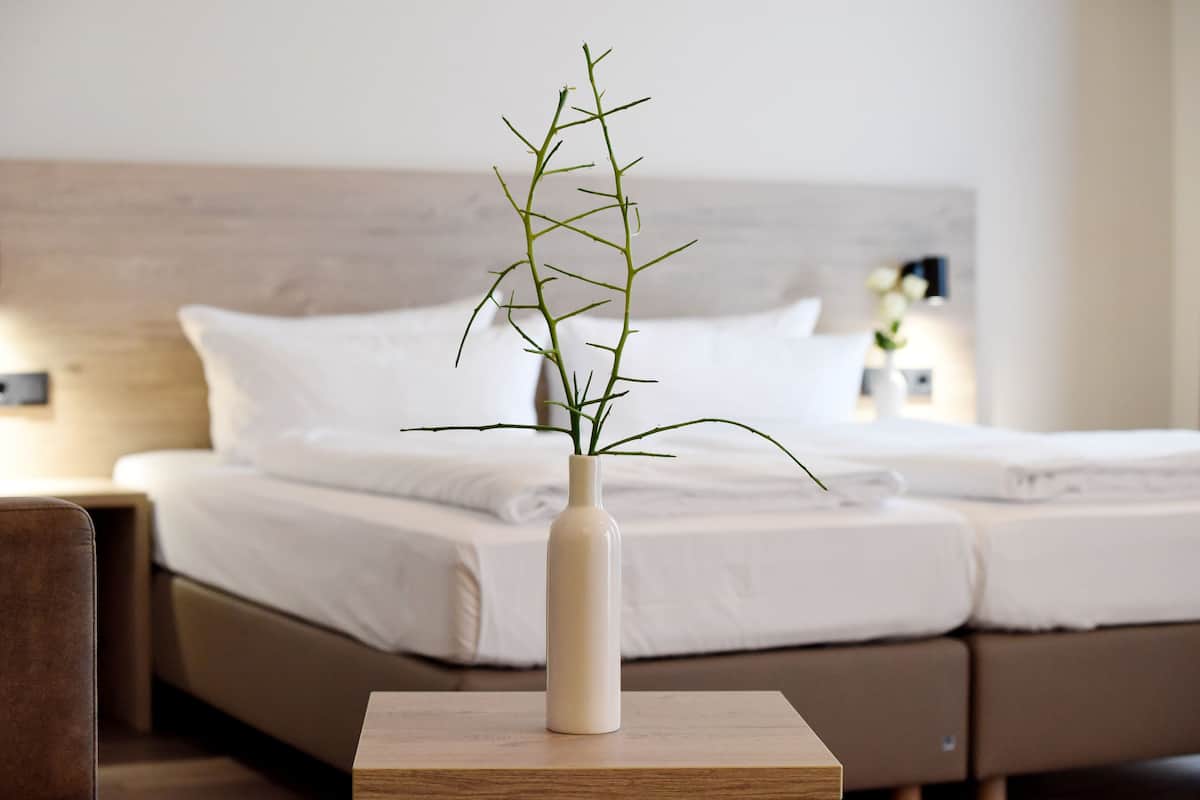 A bed with white linens, a bedside table with a vase containing a plant, and a nightstand with a lamp.