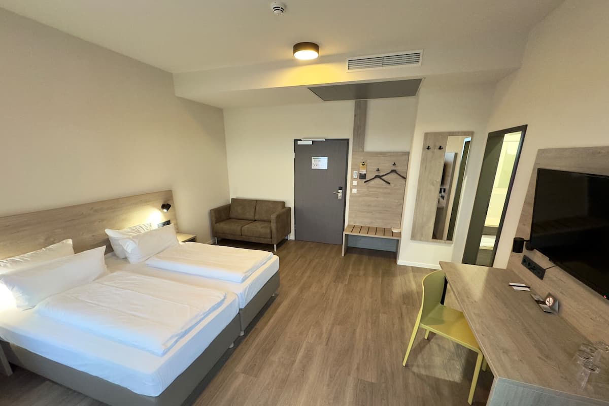 A hotel room with a bed, a desk, a chair, a TV, and a coat rack.