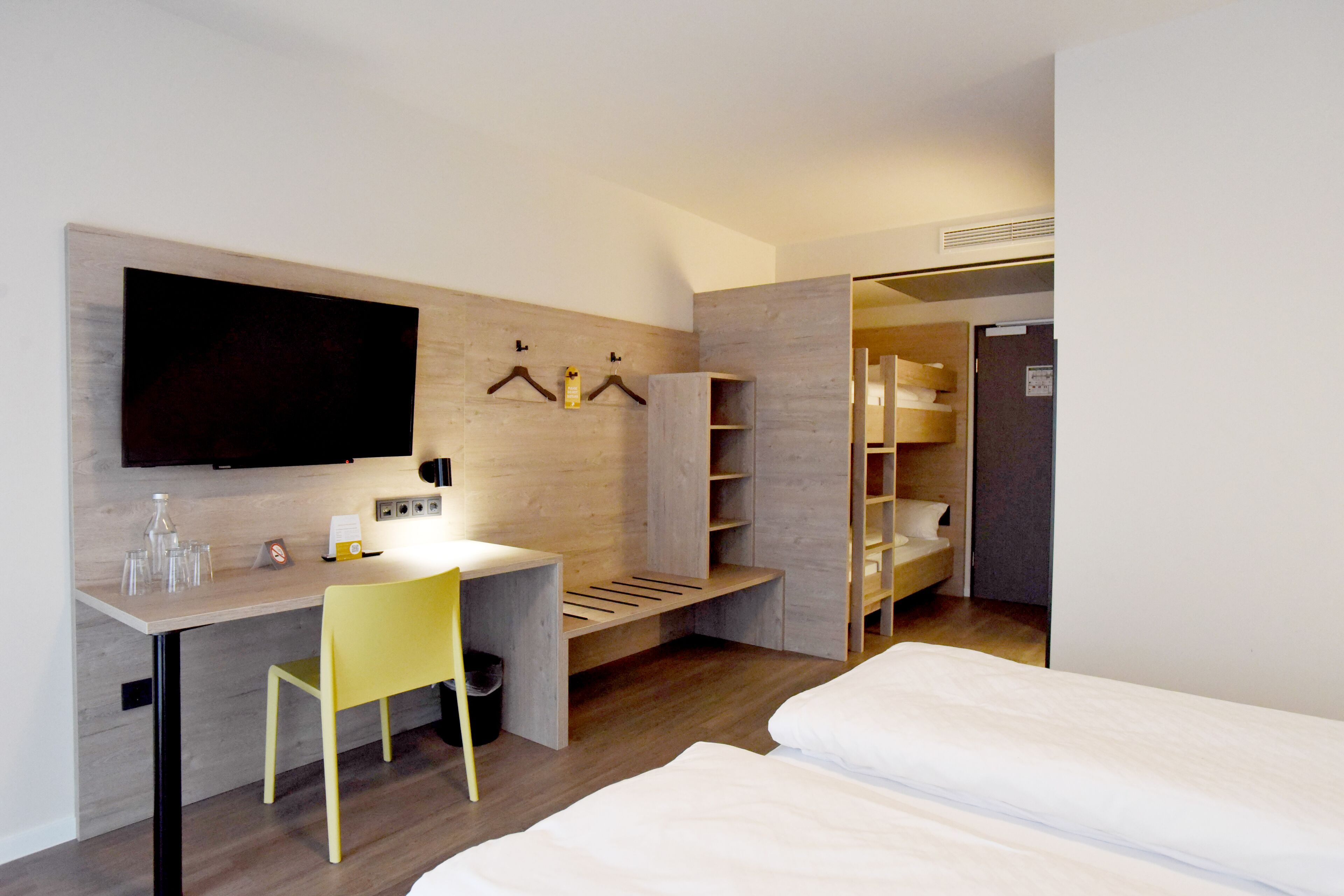 A modern hotel room with a wooden desk, a television, a bed, and a wardrobe.