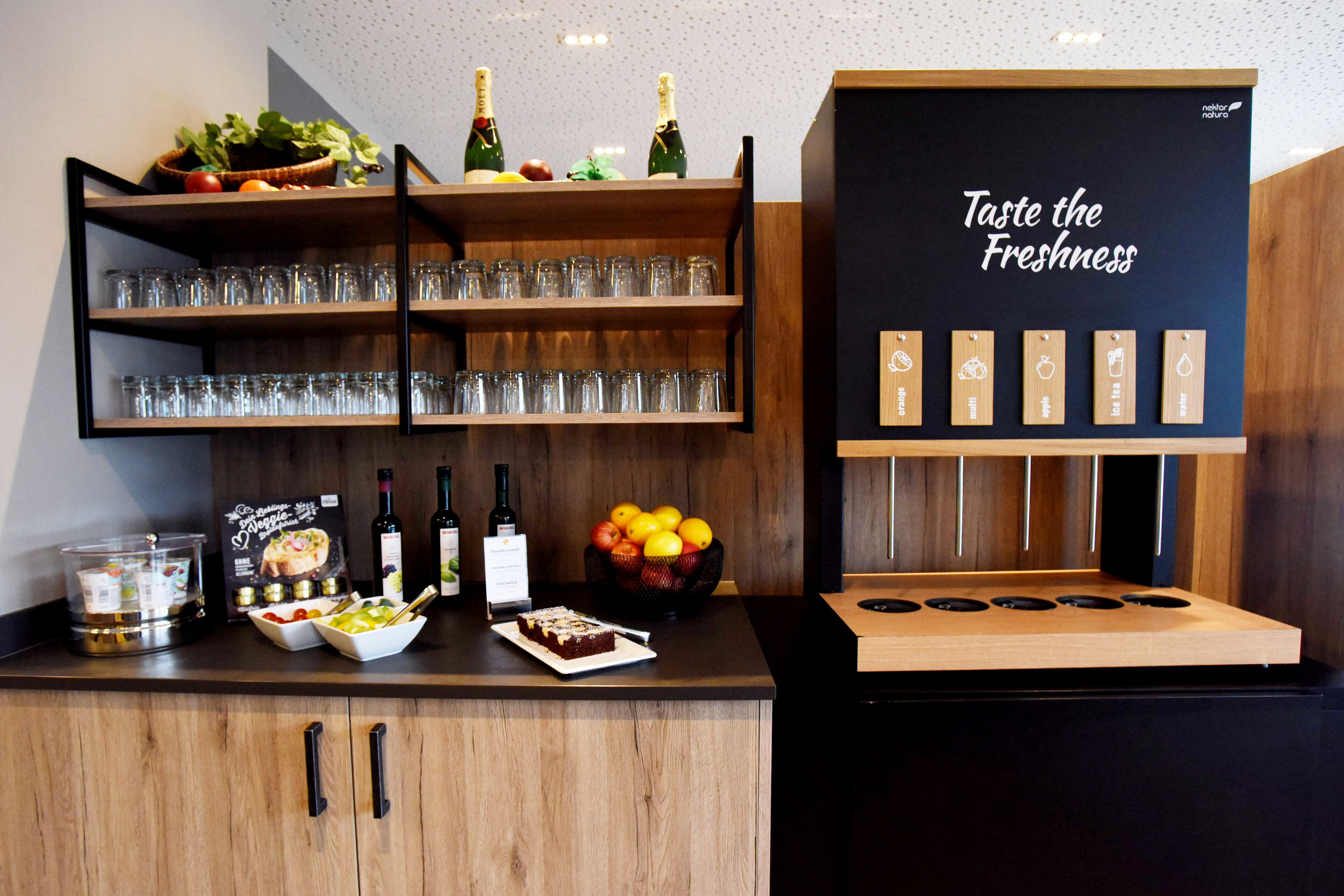A kitchen area with a wooden counter, shelves with glasses, a bowl of fruit, and a sign that says "Taste the Freshness".