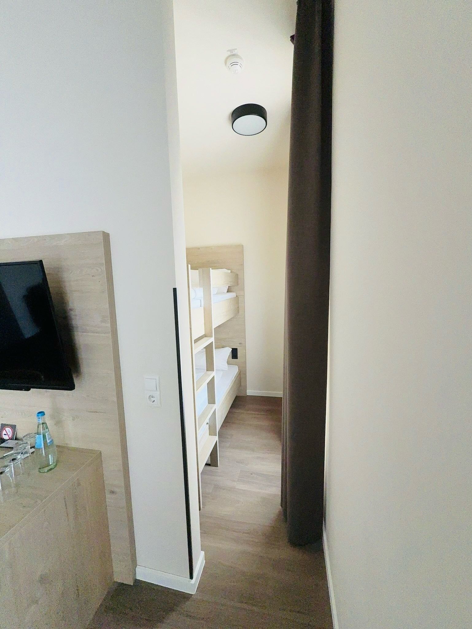 Ein schmaler Flur mit einem an der linken Wand befestigten Flachbildfernseher, einem Holzkabinett mit Flaschen und einem weißen Regal.