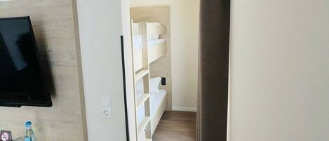 A narrow hallway with a flat-screen TV mounted on the left wall, a wooden cabinet with bottles, and a white shelving unit against the wall.