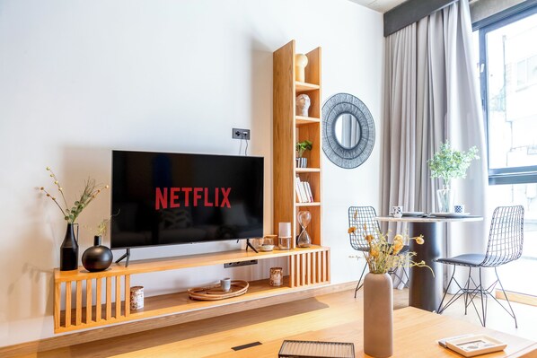 A modern living room with a large flat-screen TV displaying "NETFLIX," wooden shelving with decorative items, a dining area with a table and chairs, and a large window with curtains. - Stayhere Casablanca - Gauthier Loft Living (Casablanca)